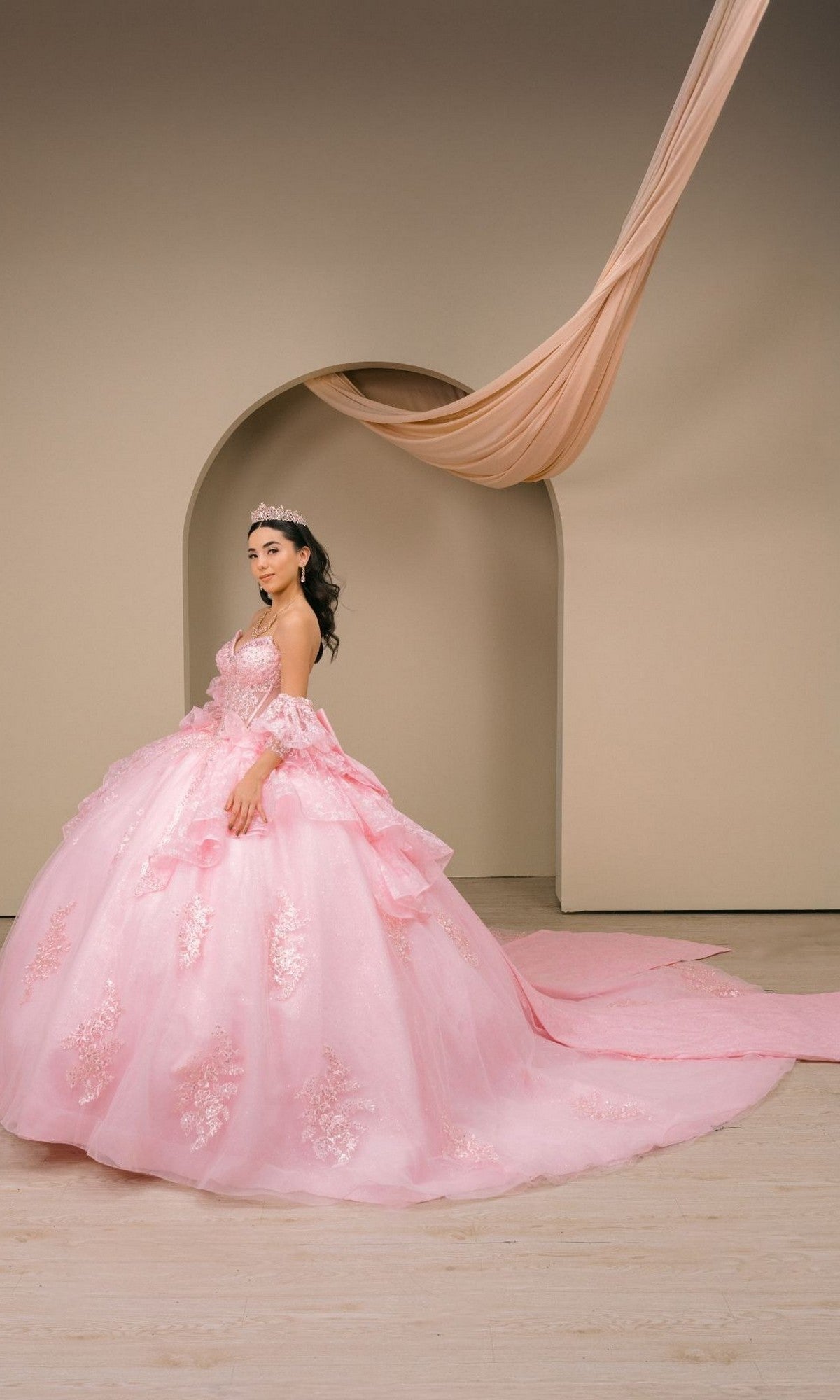 A woman in the exquisite Quinceanera Dress 8535JQ by Cinderella Couture, paired with a sparkling tiara, stands before a beige archway elegantly draped with fabric above.